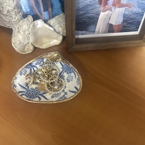 May include: A decorative seashell dish featuring a blue and white floral pattern on the interior and a gold-painted rim. The dish contains a collection of gold-toned jewelry, including hoop earrings, a crescent moon charm, and pearl-accented pieces.