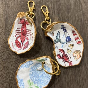 May include: Three oyster shell keychains with gold-painted edges and gold lobster clasps. One shell features a red lobster, another displays a lighthouse, crab, and seashell motif, and the third shows blue hydrangea flowers with a small white pearl charm.