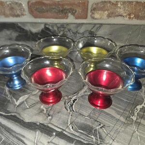 Può includere: Un set di sei piccole ciotole con piedistallo caratterizzate da gusci esterni in vetro trasparente con bordi smerlati e inserti interni metallici in tinta unita nei colori rosso, blu e giallo. Ogni ciotola poggia su una base circolare.