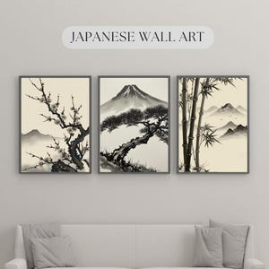 以下が含まれることがあります： 3つの額入り日本の壁アートプリント。このアートワークは、開花した枝、富士山、竹の白黒インクウォッシュペインティングを特徴としています。プリントは白いソファの上に表示されています。