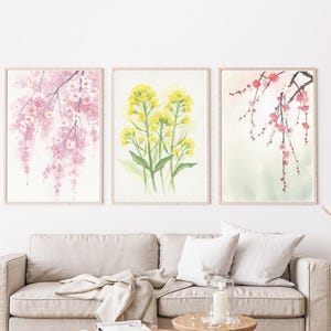以下が含まれることがあります： 3つの額入り水彩画プリント。ピンクの桜、黄色の菜の花、赤い梅の花が描かれています。ベージュのソファと木製のコーヒーテーブルの上に飾られています。