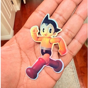 May include: Astro Boy sticker in a running pose. The sticker features a cartoon character with black hair, a yellow body, black shorts, and red boots. The sticker is held in a person's hand.