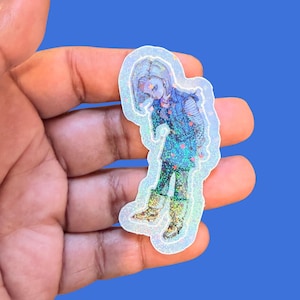 May include: A holographic sticker featuring an anime-style character with short blonde hair, wearing a blue vest, a striped long-sleeve shirt, a dark skirt, and boots. The sticker has a shimmering, glittery finish with small star-shaped accents.