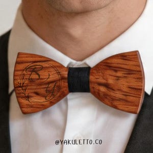 Op de afbeelding: Houten vlinderdas met een donkerbruine afwerking en een zwarte middenband. De vlinderdas heeft een cirkelvormig ontwerp met de letters "B" en "Y" en een bloemelement. De tekst "@YAKULETTO.CO" staat onderaan.