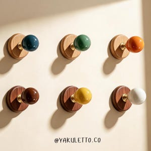 Op de afbeelding: Zes wandhaken met ronde houten basissen in lichte en donkere houttinten, verbonden door messingkleurige metalen stelen met glanzende bolvormige knoppen in blauw, groen, oranje, bruin, geel en wit. De tekst YAKULETTO.CO staat onderaan.