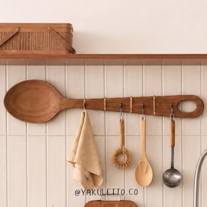 May include: A large wooden spoon-shaped wall rack with five metal hooks. Hanging from the hooks are a beige cloth, a round wooden brush, a wooden spoon, and a metal ladle. The text @YAKULETTO.CO is printed at the bottom of the wall rack display.