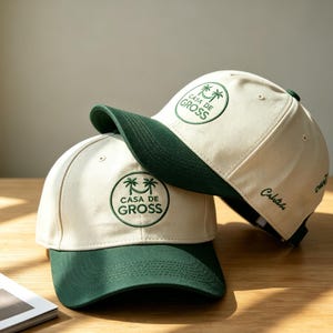 Puede incluir: Dos gorras de béisbol con corona color crema y visera verde oscuro. Cada gorra presenta un logotipo circular bordado con palmeras y el texto "CASA DE GROSS". Están sobre una superficie de madera.