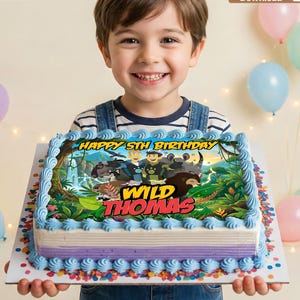 May include: A rectangular birthday cake with blue frosting and a colorful image featuring the text "HAPPY 5TH BIRTHDAY" and "WILD THOMAS". The cake is held by a smiling child, with balloons in the background.