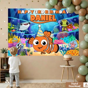 May include: A vibrant birthday banner with an underwater theme, showcasing cartoon fish, a shark, and a turtle. The text reads "HAPPY 3RD BIRTHDAY DANIEL". A child is positioned in front of the backdrop. Balloons and a small cake add to the celebratory atmosphere.