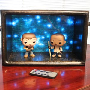 May include: A wooden display box with two character figurines and blue LED lights in the background. A remote control sits in front of the box on a red surface. The figurines are of a man with a crossbow and another with a staff.