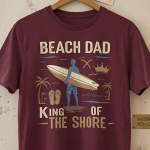 May include: Maroon t-shirt with the text "BEACH DAD KING OF THE SHORE" and a graphic of a surfer with a surfboard, palm trees, and a crown. The shirt is hanging on a wooden hanger.