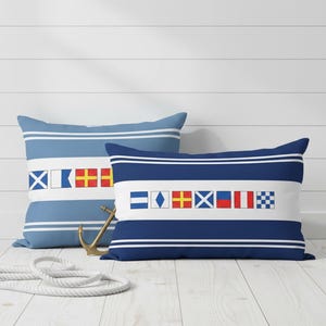 May include: Two rectangular cushions featuring nautical signal flags. One is light blue with white stripes and flags, the other navy blue with white stripes and flags. An anchor and rope are in the foreground.