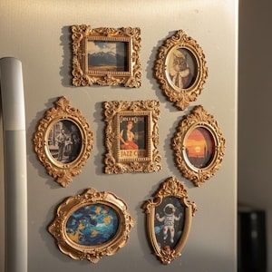 Könnte beinhalten: Eine Sammlung verzierter, goldfarbener Bilderrahmen in verschiedenen Formen und Größen, die jeweils ein einzigartiges Bild enthalten. Die Bilder zeigen Landschaften, eine Katze, ein Jazzclub-Poster und einen Astronauten. Die Rahmen sind auf einer metallischen Oberfläche angeordnet.