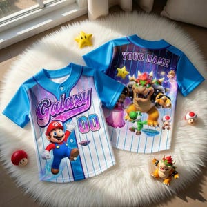 Puede incluir: Dos camisetas de b&eacute;isbol azules y blancas con gr&aacute;ficos de dibujos animados. Una camiseta presenta el texto "Galaxy" y un personaje de Mario. La otra camiseta tiene el texto "YOUR NAME" y Bowser y otros personajes. Ambas camisetas tienen mangas azules y un dise&ntilde;o de rayas blancas y azules.