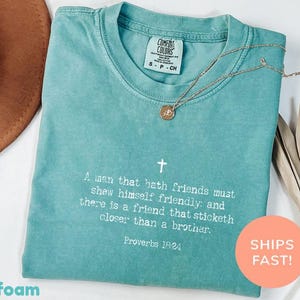 以下が含まれることがあります： シーフォームグリーンの半袖Tシャツ。胸元には白い十字架と「A man that hath friends must shew himself friendly: and there is a friend that sticketh closer than a brother. Proverbs 18:24」という白い文字がプリントされています。