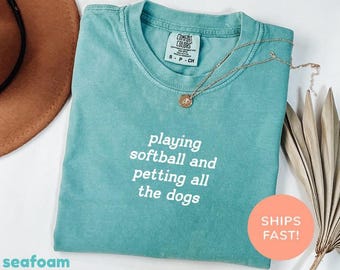 May include: Seafoam green t-shirt with the text "playing softball and petting all the dogs" in white. The shirt has a crew neck and a small tag on the collar. A gold necklace with a pendant is draped across the shirt. A brown hat is visible in the background.
