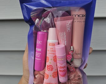 May include: A clear plastic bag filled with pink lip care products. The bag contains lip gloss, lip balm, and scrunchies. The lip products have text that reads "She", "max", "LOVE BUG", and "ULTRAMO".