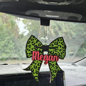 Könnte beinhalten: Ein grüner, schleifenförmiger Auto-Lufterfrischer mit schwarzen Leopardenflecken und dem Namen "Megan" in Pink. Der Lufterfrischer hängt an einer schwarzen Schnur, sichtbar im Inneren eines Autos.