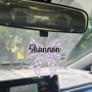 May include: A lavender-colored car air freshener shaped like a bow, hanging from a rearview mirror. The name "Shannon" is written in a cursive font across the bow. The bow has a checkered pattern.