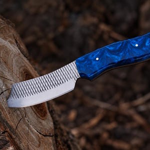 May include: A fixed-blade knife featuring a textured metal blade with a cross-hatched pattern and a smooth, polished blue handle with a marbled appearance. The handle is secured with two small metal pins and includes a metal-lined lanyard hole.