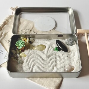 May include: A miniature zen garden kit in a rectangular metal tin. The kit includes white sand with wave patterns, small stones, a miniature succulent, a black stone, a wooden rake, and a small bottle of essential oil.