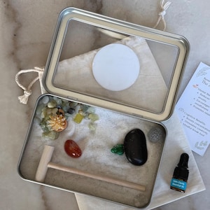 May include: A miniature Zen garden kit in a rectangular metal tin. The kit includes sand, decorative stones, a small wooden rake, and a bottle of essential oil. A card lists the benefits of the Zen garden.