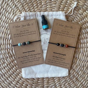 May include: Two beaded bracelets with black cords, displayed with a small essential oil bottle and a drawstring bag on a woven mat. One bracelet has turquoise and black beads, the other has red and black beads. The cards read "Wear Your Vibe."