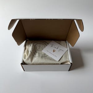 May include: A white box with a brown interior, lid open. Inside, a beige cloth bag and a small white card with "Thank you" and a heart symbol are visible. The box contains a product from "The Flourish Box".