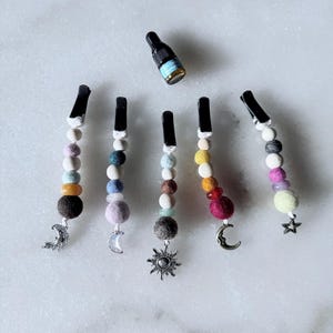 Op de afbeelding: Een verzameling kleurrijke haarclips met kralen en hemelse bedels, waaronder manen en sterren. Elke clip heeft een unieke combinatie van vilten ballen en kralen in verschillende kleuren. Er is ook een kleine fles essentiële olie aanwezig.