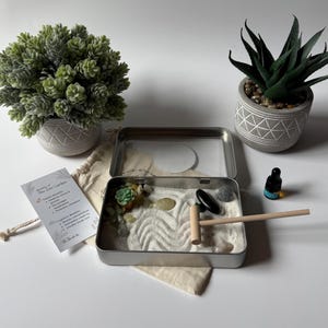 May include: A miniature Zen garden kit with a metal tray, sand, rocks, and a small rake. The kit includes a small succulent, a bottle of essential oil, and two decorative potted plants. A card lists the benefits of the Zen garden.