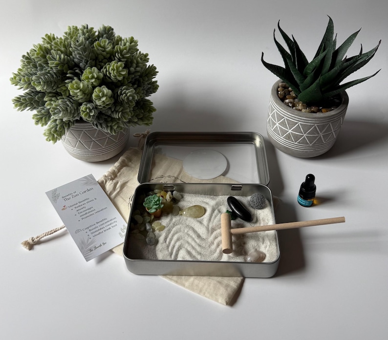 May include: A miniature Zen garden kit with a metal tray, sand, rocks, and a small rake. The kit includes a small succulent, a bottle of essential oil, and two decorative potted plants. A card lists the benefits of the Zen garden.