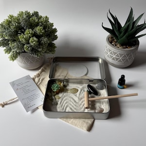 May include: A miniature Zen garden kit with a metal tray, sand, rocks, and a small rake. The kit includes a small succulent, a bottle of essential oil, and a small plant in a decorative pot. A small card reads "Benefits of The Zen Garden".