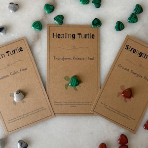 Puede incluir: Tres tarjetas de papel kraft con los textos "Dream Turtle", "Healing Turtle" y "Strength Turtle". Cada tarjeta tiene una pequeña piedra en forma de tortuga adjunta. Piedras en forma de corazón de varios colores rodean las tarjetas.