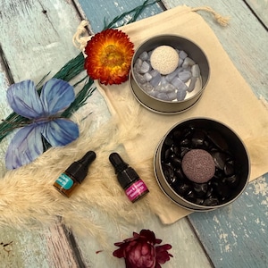 May include: An overhead shot of a wellness set featuring two small tins filled with stones and a porous ball, two essential oil bottles, a drawstring bag, and dried flowers. The tins contain blue and black stones. The essential oil bottles have labels.