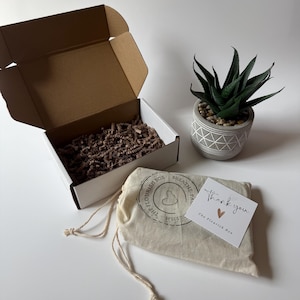 May include: A white box with a brown interior and an open lid, a small potted succulent, and a drawstring bag with a printed logo. A card with "Thank you" and a heart is next to the bag.