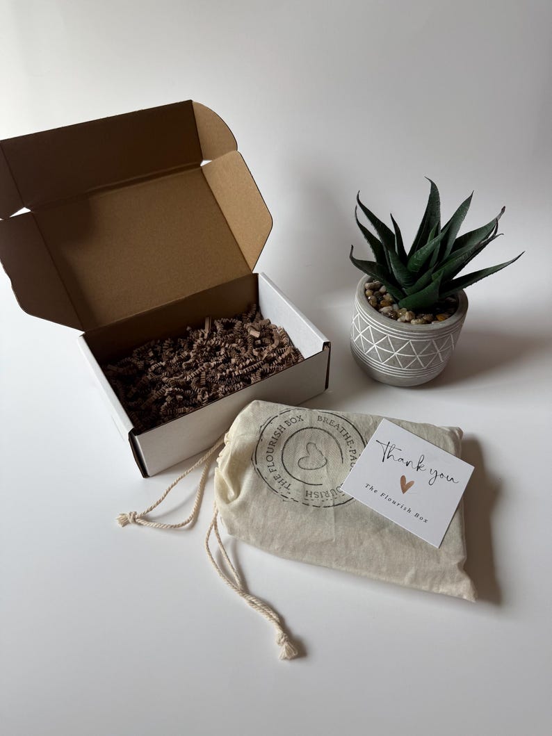 May include: A white box with a brown interior and an open lid, a small potted succulent, and a drawstring bag with a printed logo. A card with "Thank you" and a heart is next to the bag.