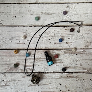 May include: A black necklace with a filigree pendant and a small essential oil bottle with a blue label. Colorful wooden beads in various sizes and colors are scattered around the necklace on a distressed white wooden surface.