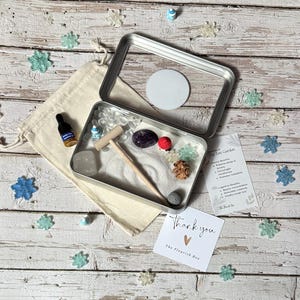 May include: A small, open metal box containing a miniature zen garden with white sand, a wooden rake, and decorative stones. A small bottle, a drawstring bag, and a card that says "thank you" are also present.