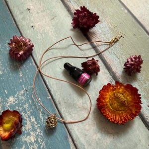 Puede incluir: Un collar con colgante dorado con un diseño calado redondo, exhibido con una pequeña botella de aceite esencial y flores secas en tonos rojos y naranjas. El collar tiene un cordón beige y un cierre dorado.