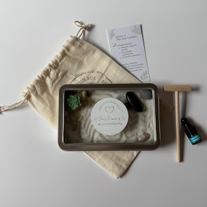 May include: A Zen garden kit featuring a metal tray, sand, decorative stones, a small wooden rake, a drawstring bag, and a bottle of essential oil. The kit includes a card detailing the benefits of a Zen garden.
