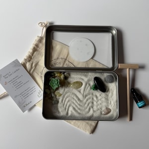 May include: A miniature Zen garden kit in a metal tin, with sand, decorative stones, and a small rake. Includes a small bottle and a card listing the benefits of the Zen Garden. The kit is designed for relaxation and mindfulness.