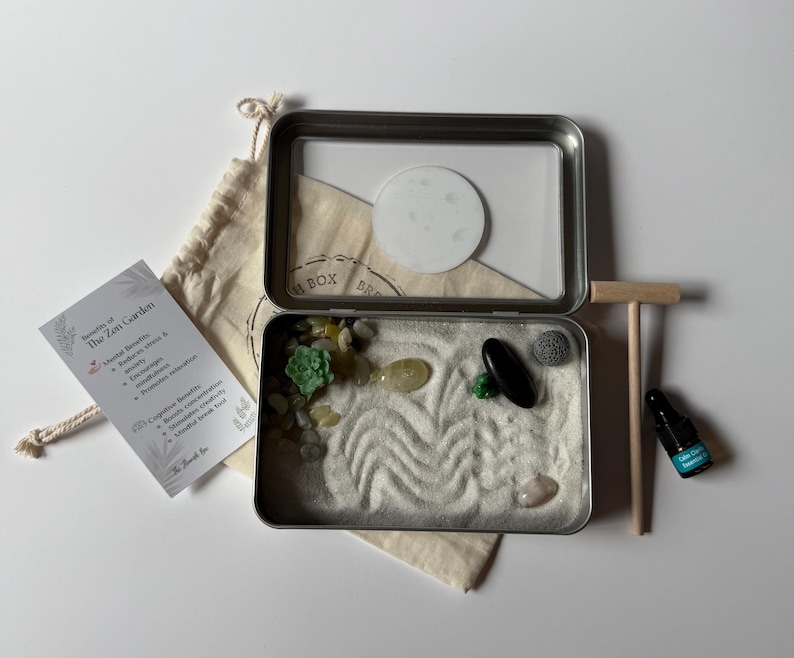 May include: A miniature Zen garden kit in a metal tin, with sand, decorative stones, and a small rake. Includes a small bottle and a card listing the benefits of the Zen Garden. The kit is designed for relaxation and mindfulness.