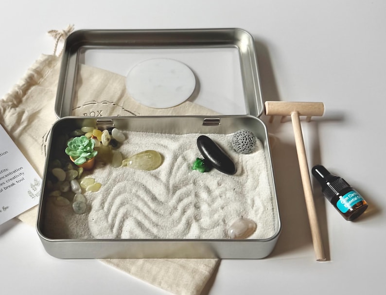 May include: A miniature zen garden kit in a rectangular metal tin. The kit includes white sand with wave patterns, small stones, a miniature succulent, a black stone, a wooden rake, and a small bottle of essential oil.