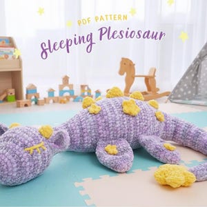 Könnte beinhalten: Ein handgefertigtes, plüschiges, lila Plesiosaurier-Spielzeug mit gelben Akzenten. Das Spielzeug liegt auf einer Spielmatte, darüber steht der Text "Sleeping Plesiosaur". Das Spielzeug hat ein weiches, strukturiertes Aussehen.
