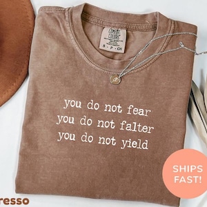 May include: Espresso-colored t-shirt with the text "you do not fear, you do not falter, you do not yield" in white. The shirt has a crew neck and a small tag on the collar. A silver necklace with a pendant is draped across the shirt.