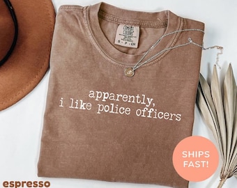May include: A brown short-sleeved t-shirt with a crew neckline featuring the white printed text 'apparently, i like police officers' on the chest. The garment has a Comfort Colors brand label inside the collar indicating a size small.