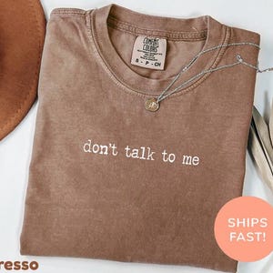 Puede incluir: Camiseta color espresso con la frase "don't talk to me" en letras blancas. La camiseta tiene cuello redondo y una pequeña etiqueta en la parte superior de la espalda. Un collar plateado con un colgante descansa sobre la camiseta. Una pegatina redonda con la frase "SHIPS FAST!" es visible.