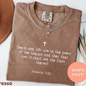 May include: A brown short-sleeved t-shirt featuring a white cross and the text Death and life are in the power of the tongue: and they that love it shall eat the fruit thereof. Proverbs 18:21 printed on the front with a Comfort Colors label.