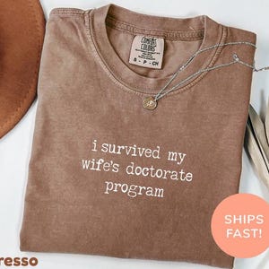 May include: Espresso-coloured t-shirt with the text "i survived my wife's doctorate program" in white. The shirt has a crew neck and short sleeves. A silver necklace with a pendant is draped across the shirt.