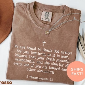 May include: A brown short-sleeved t-shirt featuring a white cross and the text: We are bound to thank God always for you, brethren, as it is meet, because that your faith groweth exceedingly, and the charity of every one of you all toward each other aboundeth; 2 Thessalonians 1:3.